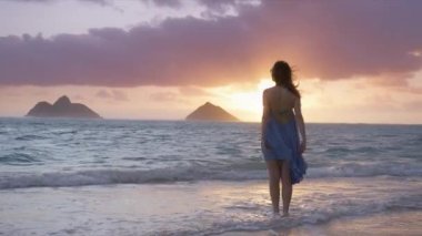 Okyanus esintisi elbisesiyle uçan, altın gündoğumunda ya da günbatımında Lanikai sahilinde dalgaların arasında duran mutlu, rahat kadının yavaş arkası görüntüsü. Hawaii Yaz Tatili Kadın Turist