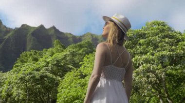 Genç seksi kadın, egzotik zümrüt yeşili Pasifik adasında beyaz boho moda elbisesi ve şık hasır şapkasıyla Hawaii 'de güneşli yaz yolculuğunda eski volkan dağlarının yeşil tepelerine bakıyor.
