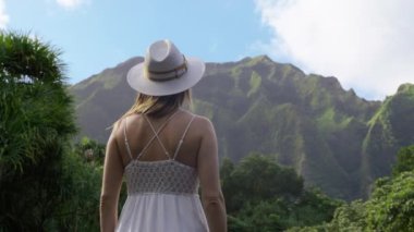 Genç seksi kadın Hawaii 'de güneşli yaz yolculuğunda eski volkan dağının yeşil tepelerine bakıyor. Egzotik yeşil Pasifik adasında gezgin. Beyaz boho elbisesi ve şık hasır şapkasıyla Oahu.