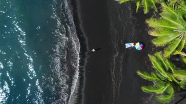 Tropik Hawaii adasında bir kadın. Havadan mutlu turist egzotik siyah lav kumsalında güneşlenirken okyanus esintisinde sallanan güzel yeşil palmiyeler manzaralı mavi deniz dalgaları 4K USA