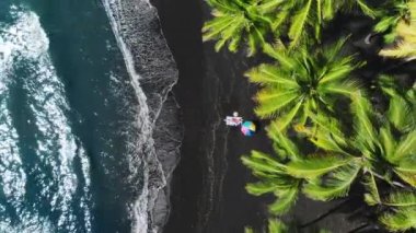 Seksi kırmızı mayo giymiş, egzotik siyah lav kumlu Hawaii plajında güneşlenen mutlu bir kadın. Sinema salonunun tepesinde güzel yeşil palmiyeler okyanus esintisinde sallanıyor mavi okyanus dalgaları kıyıda 4K