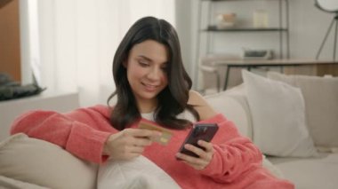 Modern evdeki kanepede oturan akıllı telefon ve kredi kartı tutan gülümseyen genç bir kadın. Anında kolay ödemeler yapan mutlu bayan alışverişçi, online alışveriş yapan müşteri, e bankacılık uygulaması