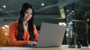 Kendine güvenen Asyalı iş kadını güzel gülüşü olan dizüstü bilgisayara bakıyor, ağır çekimde. Mutlu başlangıçlı girişimci dizüstü bilgisayarına e-posta yazıyor, açık uzay dairesindeki masada oturuyor.