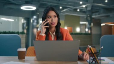 Gülümseyen Asyalı iş kadını dizüstü bilgisayarla telefonda konuşuyor ofis masası 4K 'da oturuyor. Mutlu karışık ırk bayan yöneticisi mobil arama yapıyor proje detaylarını, ticaret anlaşmasını, iş anlaşmasını onaylıyor
