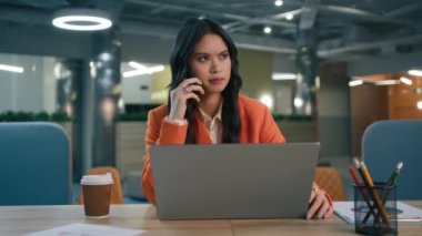 Endişeli karışık ırk bayan yöneticisi mobil arama yapıyor proje detaylarını, ticaret anlaşmasını, iş anlaşmasını onaylıyor. Yoğunlaşmış Asyalı genç iş kadını ofis masasında dizüstü bilgisayarla konuşuyor.