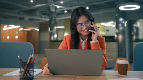 若いアジア系アメリカ人のビジネスレディーがオフィスで成功した仕事を楽しんでいます 机の上でノートパソコンを使いスマートフォンで話をする民族の女性良いニュースを祝う楽しみを持っている — ストック動画