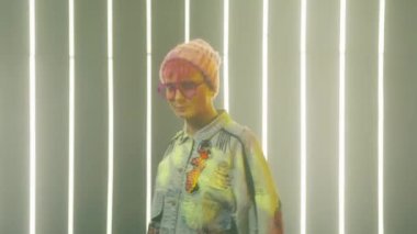 Pembe saçlı cesur kızın kulüpteki portresi. Pembe çerçeveli moda gözlüğü takmış ukala, küstah bir kadın ve bol bol kot pantolon giyip şeker çiğniyor. 20 'li yaşlarda bir kadın kameraya yavaş çekimde bakıyor.