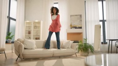 Modern koltukta mutlu bir şekilde zıplayan renkli, heyecanlı genç bir kadın. Afro kız ev sahibi gün içinde taşınırken eğleniyor, ipotek onayını kutluyor, yeni bir daireye taşınıyor. Banka ipoteği kavramı