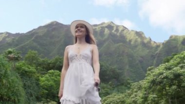 Renkli, zarif beyaz şapkalı ve boho elbiseli genç, sarışın bir kadın. Hareketli arka planda yüksek yeşil Hawaii dağları manzarası olan RED kamerasında yavaş çekim yapıyor. Oahu adası gezisi