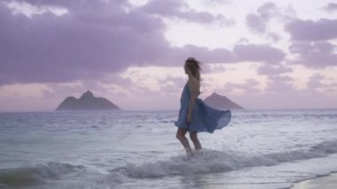 Hawaii adasında yaz tatili, seyahat geçmişi. Lanikai plajında Mokulua adaları manzaralı mutlu bir kadının mavi okyanusa yürüyüşü. Sinema rüzgarlı gündoğumu, gökyüzünde pembe mor bulutlar.