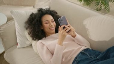 Rahat Afro-Amerikan kadın elinde akıllı bir telefonla kanepede oturup sosyal medyanın rahatlamasını izliyor. Akıllı telefon teknolojisi kullanan kıvırcık kadın, yavaş çekimde internetten alışveriş yapan genç kız 4K