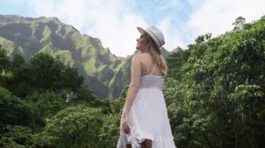 Yazın güneşli bir günde yeşil parkta yürüyen çekici, gülümseyen esmer kadın. Havai adasında moda şapkalı ve elbiseli mutlu bir kadın doğa hayranı. Yavaş çekim 4K Kırmızı Kamera Videosu