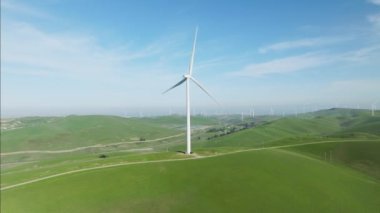Çevre dostu kavram. Güneşli bir günde, arka planda parlak yeşil tepeler bulunan rüzgar türbininin manzaralı görüntüsü. Rüzgar türbini, sürdürülebilir elektrik üretmek için dönen büyük döner bıçaklar.