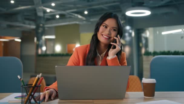 有道德的女人坐在办公桌前 打开笔记本电脑 用智能手机交谈 开心地庆祝好消息慢下来 兴奋的年轻亚裔美国女商人在办公室里享受着成功的工作计划 — 图库视频影像