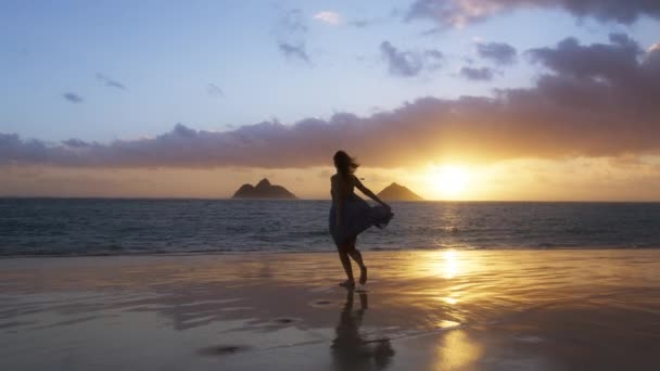 シルエット幸せな陽気な女の子の水の砂のビーチで夏の日の出を実行している 海の波で回転するドレスを飛んでスリム女性 陽気な女性は海岸で楽しみを持っています美しいハワイの自然を楽しむ — ストック動画