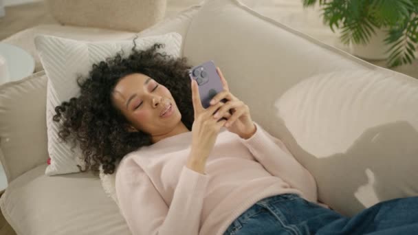 アフリカ系アメリカ人の女性が太陽の光の下で家にスマートフォンを持って笑っている 快適なソファの上に横たわるソーシャルメディアを見て色の女性 中の女の子の肖像 スローモーションでのスマートフォンエンターテイメント — ストック動画