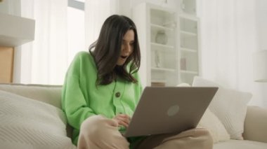 Heyecanlı genç kadın dizüstü bilgisayara bakıyor ve online başarıyı kutluyor. Öforik kadın evdeki kanepede oturarak yeni bir iş fırsatı elde ediyor. Motive olmuş kız e-postada iyi haberler okuyor, zaferden dolayı mutlu.