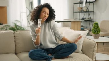 Güzel kıvırcık saçlı, dans eden, saç fırçasında şarkı söyleyen, dairede eğlenen mutlu Afro-Amerikan bayanın ağır çekimleri. Kanepede oturan komik renkli kadın. İnsan yaşam tarzı ve eğlence kavramı