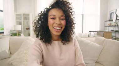 Genç, Afro-Amerikan melez, video blogcu kadın el sallıyor, webcam 'e bakıyor, hipster kız kamerayla konuşuyor video konferans görüşmesinde kanepede oturuyor. Sosyal uzaklıkla buluşan sanal sohbet arkadaşları