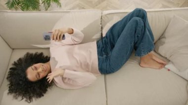 Evdeki rahat oturma odasında akıllı telefon kullanan mutlu, güzel Afro-Amerikan kadın. Kadın rahat bir kanepede dinleniyor. Renkli kadın internette geziniyor ve selfie resmi sosyal medya adı verilen video yapıyor.