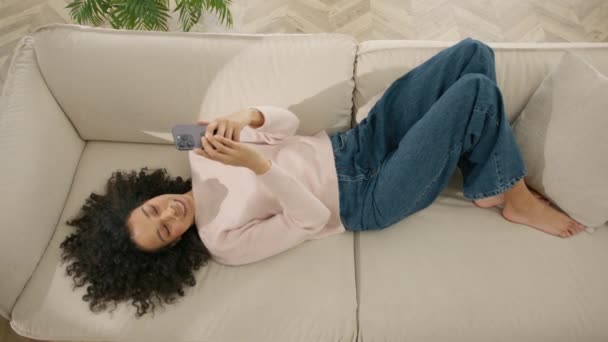 色彩艳丽的女人上网做视频自选图片社交媒体 快乐美丽的非洲裔美国女性在舒适的客厅里用智能手机在家里 女人躺在舒适的沙发上 — 图库视频影像