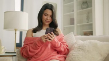 Akıllı telefon kullanan, kanepede oturan neşeli genç bir kadının sinematik çekimi. Modern evde cep telefonu uygulaması kullanan gülümseyen bir kadın. Güzel kız kameraya bakarken dinleniyor, telefona mesaj atıyor.