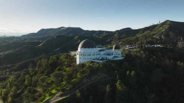 カリフォルニア州ロサンゼルス2023年3月 ドーム建築の周りを飛ぶグリフィス天文台 日没の明るい緑の丘の上にハリウッドの看板 緑の春公園の空中景観ランドマーク建物 — ストック動画