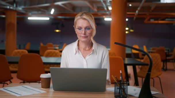 带着眼镜的疲倦的女老板在现代阁楼风格的办公室工作 背景是运动的 成熟迷人的女人工作到很晚 50岁的女商人在笔记本电脑上输入电子邮件 喝咖啡 — 图库视频影像