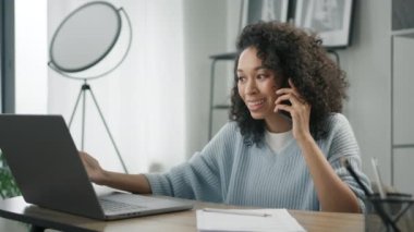 Heyecanlı genç Afrikalı Amerikalı iş kadını evde serbest çalışmanın tadını çıkarıyor. Renkli bir kadın dizüstü bilgisayara bakıyor, akıllı telefondan konuşuyor, iyi haberleri ağırdan alarak kutluyor.