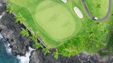 Golf arabası parlak yeşil golf sahasında mükemmel bir patikadan geçiyor. Yeşil golf kulübünün tepesinden aşağı 4K. Manzaralı hava manzarası dramatik siyah lav kayalık kıyıları, Büyük Ada tropikal kıyısındaki mavi okyanus Hawaii