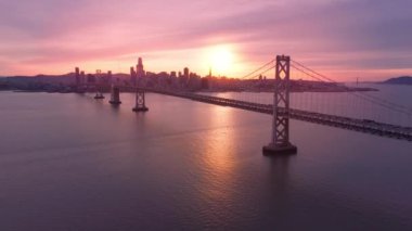 Körfez köprüsü inşaatı boyunca su yüzeyinde uçan insansız hava aracı. 4K şehir manzarası. San Francisco şehir merkezinde gökdelenleri olan Bay Bridge 'in güzel hava manzarası sinemada altın gün batımı ışığı.
