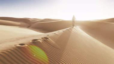 4K hava aracı, çölün ortasında güneşten parlayan altın mercekle yürüyen turist bir kadını görüntülüyor. Arka planda altın gün batımında kum tepesinde yürüyen gezgin kadının yavaş hareketi
