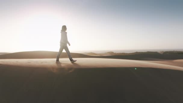 砂漠の風の強い日に白人女性に続くドローン レンズフレアと風光明媚な黄金の夕日の丘の上に幸せなハイカーの女性 無料の女性旅行者が映画の自然景観で歩く — ストック動画
