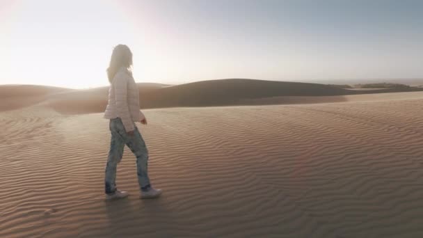 レンズフレアを持つ風光明媚な黄金の夕日の砂丘の上に冒険ハイカー 若い女性旅行者の映画的な自然の風景の中を歩く 風の強い日に白人女性に続く4Kドローン — ストック動画