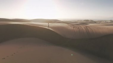 Kumlu tepenin tepesindeki uzak yürüyüşçü manzaralı altın gün batımında mercek parlamasıyla. Genç bayan gezgin sinematik doğal manzaranın önünden geçiyor. 4K İHA rüzgarlı bir günde Kafkasyalı kadını takip ediyor