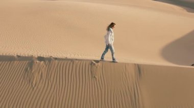 Yavaş çekim 4K hava aracı görüntüleri gezgin kadının altın günbatımında kum tepeciğinde ayak izleri bıraktığını gösteriyor. Kaliforniya 'nın altın kumu üzerinde uzun gölgeler. Çölde doğa yürüyüşü yapan turist kadın.