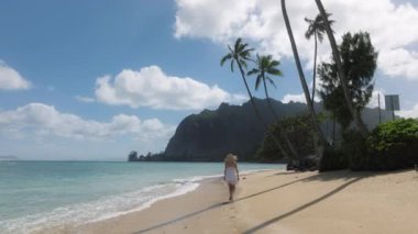 Yavaş çekim yapan kadın tropikal kumsalda yürüyor ve ayak izleri bırakıyor. Cennet adası Oahu 'da yaz tatilindeki kadın turist. Beyaz mini boho elbiseli kadın yeşil Hawaii adalarında, ABD 4K
