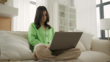 Uzaktan eğitim alan bir sınıf modern apartman dairesinde oturuyor. Genç esmer kadın üniversite öğrencisi dizüstü bilgisayarla öğreniyor, notlar yazıyor, webinar veya sanal eğitim izliyor.