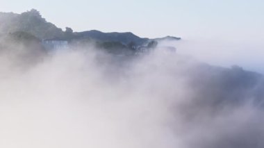 İnsansız hava aracı, güneş doğarken bulutlarla kaplı dağ tepesindeki malikanenin etrafında uçuyor. Yeşil ormanın etrafında süzülen sinematik bulutlar dağ zirvesinde mavi gökyüzü arka planda 4K