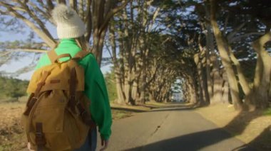 Genç milenyum gezgini ya da turist sırt çantalı seyahat blogcusu güzel selvi ağacı tünelinde yürüyor. Kuzey Kaliforniya doğa parkı konsepti 4K seyahat eder. Kamera bir kadını gezide ya da macerada takip eder