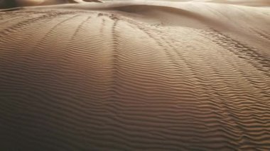İnsansız hava aracı, güçlü rüzgarın yarattığı dalgalı yüzeyi ile altın kumun üzerinde alçaktan uçuyor. Derin gölgeli kum tepesi dokusu. Gün batımında hava manzaralı çöl doğası 4K. Güzel Sahra Çölü manzarası