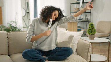 Uçan kıvırcık saçlı mutlu Afro-Amerikalı kadın evde şarkı söylüyor, başarılı yaşam tarzını kutluyor, kanepede dans ediyor, hafta sonları modern oturma odasında karaoke müzik eğleniyor.