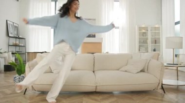 Latin kız gülüyor, rahat koltukta dinleniyor, çok mutlu hissediyor, evden eve taşınmayı kutluyor, daire kiralıyor. Mobilya tasarımı. Mutlu Latin Amerikalı kadın modern yumuşak kanepeye atlıyor 4K