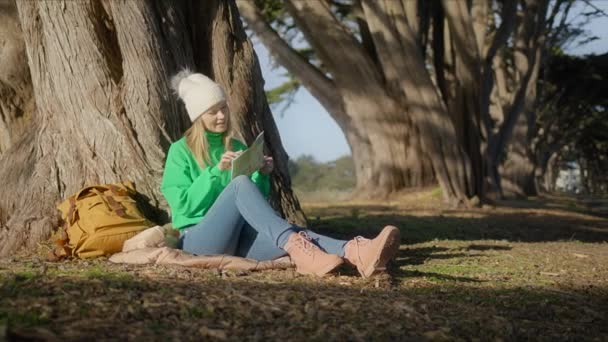 ハッピーヒップスター女性紙の道路地図を見ながら地面ヒノキの木にもたれに座って 笑顔で ときに明るい太陽を見て辞退 ハイキングマップを見て森の中でロードトリップを計画 — ストック動画