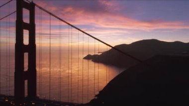 Golden Gate Köprüsü 'nün arka planında gül sarısı bulutlarla destansı günbatımı gökyüzü. Pembe gökyüzünde gece aydınlatmalı asma köprünün hava görüntüleri. San Francisco tarihi sinemada gün batımı 4K