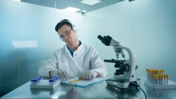 在显微镜下观察满意的科学家男性画像 分析药物实验室无线耳机听音乐时的培养皿样品 医疗实验室的人 — 图库视频影像