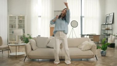 Kulaklıklı mutlu Latin Amerikalı kadın evde şarkı söylüyor, başarılı bir yaşam tarzını kutluyor, koltukta dans ediyor, hafta sonları modern oturma odasında karaoke müzik eğlencesinin tadını çıkarıyor.