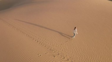 Beyaz kadın üzerinde altın ışıkta uçan drone. Sinematik kum tepeleri çölünde gezen maceraperest. Gün batımında sinemanın önünden geçen kadın gezginin yukarıdan görünüşü 4K