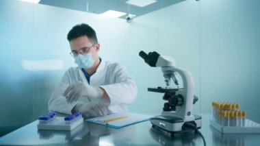 Erkek bilim adamı virüs örnekleriyle test tüplerine bakıyor ve modern araştırma laboratuvarında notlar alıyor. Koruyucu tıbbi maske takan bir bilim adamı. İlerici bilimsel biyoteknoloji tıp laboratuvarı