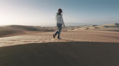 Çölde rüzgarlı bir günde turist kadının arkasında uçan drone. Özgür kadın gezgin sinematik doğal manzara 4K 'nın yanından geçiyor. Kum tepesinin tepesinde mutlu yürüyüşçü kadın mercekli altın gün batımında